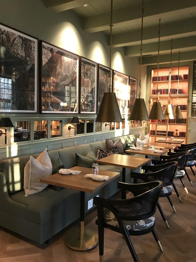 Gemütlicher Gastronomiebereich eines Hotels mit langer salbeigrüner Sitzbank, Holztischen, schwarzen Armlehnenstühlen, konischen Messing-Hängeleuchten und Rahmenbildern an der Wand