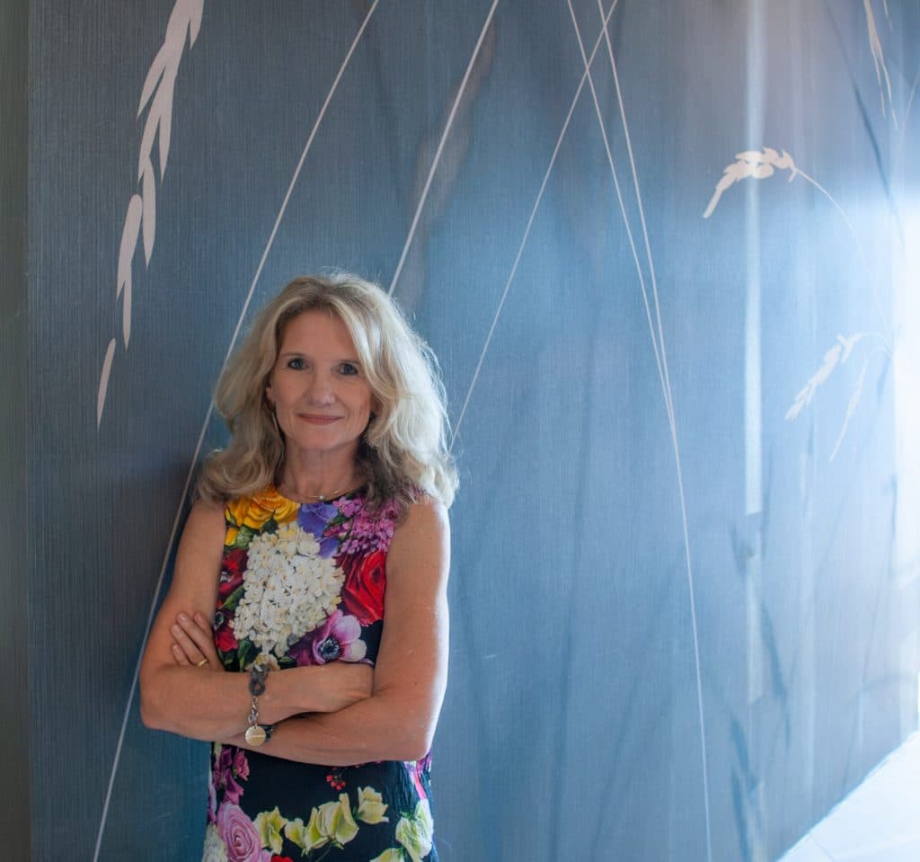 Interior Designerin Susanne Wefers im gemusterten Kleide steht vor einer stilvollen bläulichen Motivtapete mit feinen Linien und Vogelmotiven